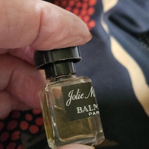 May include: A small, rectangular perfume bottle with a black cap. The label reads "Jolie M BALM PAR". The bottle contains a light amber liquid. The bottle is held between fingers.