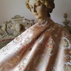 May include: A pale peach-coloured fabric featuring an intricate woven pattern of white, red, and green floral motifs and swirling scrollwork. The textile is draped over the shoulders of a sculpted bust of a person with wavy hair and neutral features.