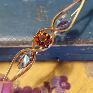May include: A gold-toned brooch with three faceted stones. The center stone is a round, orange-red color. The two outer stones are oval-shaped and have a blue-green iridescent finish.