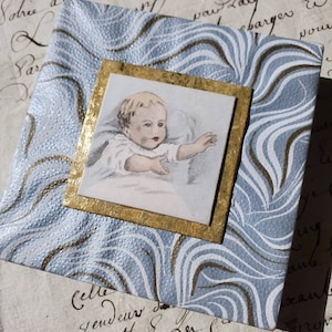 May include: A blue and gold decorative box with a framed image of a baby. The image is in a vintage style and the baby is wearing a white gown.