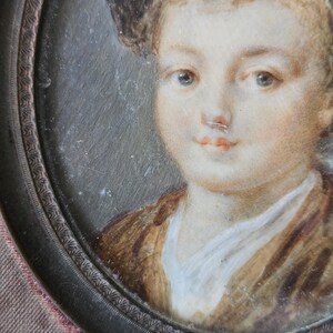 May include: Oval portrait miniature of a young person. The subject has fair skin, blue eyes, and light brown hair. They are wearing a brown jacket over a white shirt. The portrait is set in a dark metal frame with a decorative edge.