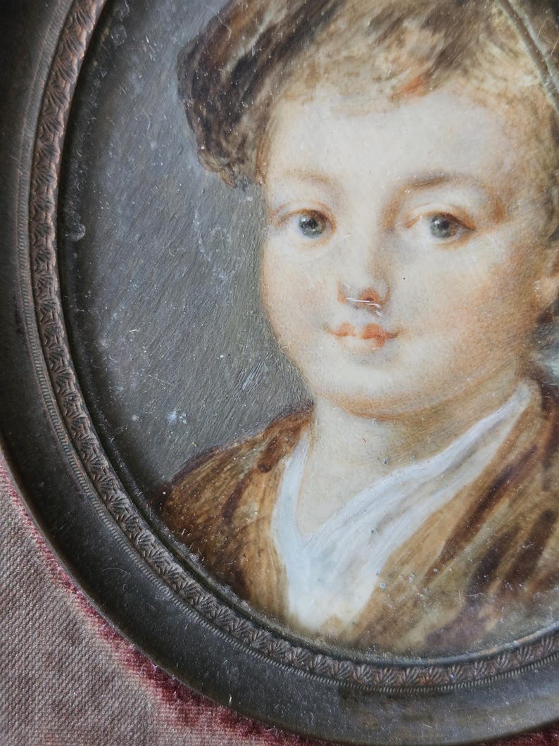 May include: Oval portrait miniature of a young person. The subject has fair skin, blue eyes, and light brown hair. They are wearing a brown jacket over a white shirt. The portrait is set in a dark metal frame with a decorative edge.