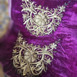 Puede incluir: Una tela de terciopelo morado intenso con dos motivos florales bordados en tono dorado idénticos. Cada diseño incluye un intrincado trabajo de hilo metálico, patrones de hojas y un grupo central de pequeñas cuentas redondas color crema.