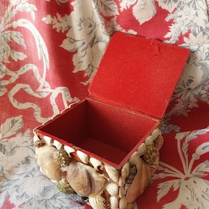 Puede incluir: Una pequeña caja de madera cubierta de conchas marinas. La caja está forrada de terciopelo rojo y tiene una tapa con bisagras.