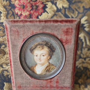 May include: A small, antique portrait of a child in a velvet-covered frame. The portrait is a miniature painting, featuring a young person with a brown jacket and a white collar. The frame is a muted red color.