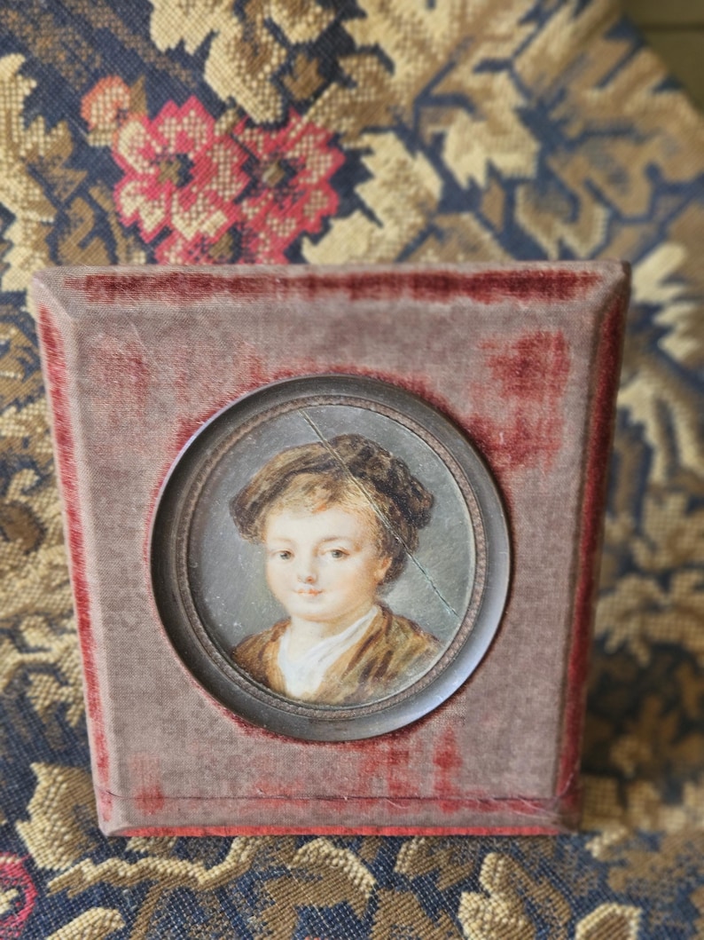 May include: A small, antique portrait of a child in a velvet-covered frame. The portrait is a miniature painting, featuring a young person with a brown jacket and a white collar. The frame is a muted red color.