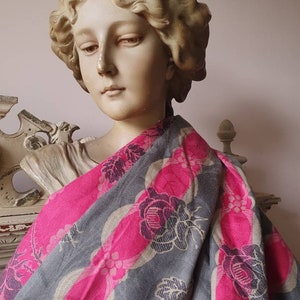 Op de afbeelding: Een klassieke buste gedrapeerd met een grijze en roze bloemenpatroon sjaal. De sjaal heeft een herhalend ontwerp van roze ovalen en grijze bloemmotieven. De buste is lichtbeige van kleur en heeft gedetailleerde gelaatstrekken.