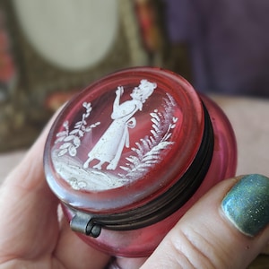 Puede incluir: Pequeña y redonda caja de baratijas de cristal rosa antiguo con tapa abisagrada. La tapa presenta un camafeo blanco de una mujer y detalles florales. La caja tiene un cierre metálico y un borde oscuro. La caja se sostiene en una mano.