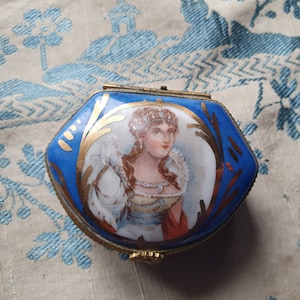 May include: A small, hexagonal trinket box with a blue enamel exterior and a portrait of a woman. The portrait is framed by gold accents. The box has a gold-colored metal frame and clasp.