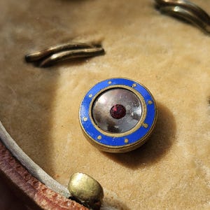May include: A close-up of a round, blue and gold button with a red jewel in the center. The button is surrounded by a white border and a ring of gold dots.