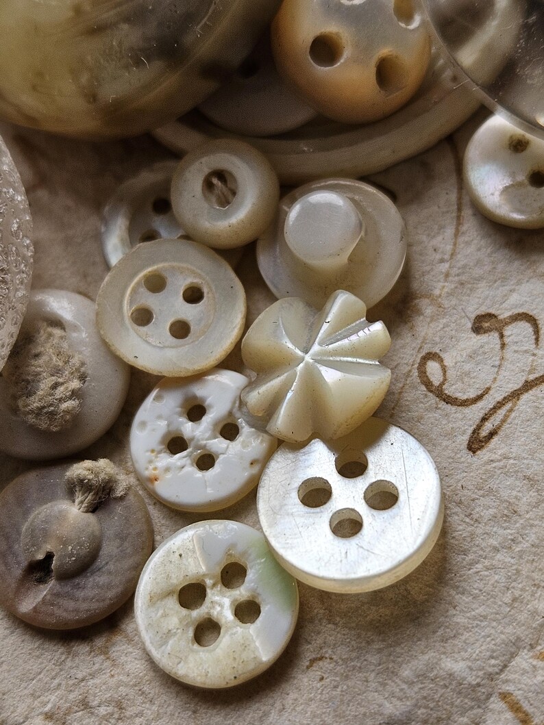 May include: A collection of white and off-white buttons with various designs and sizes. Some buttons have four holes, while others have a single hole. The buttons are arranged on a white surface.