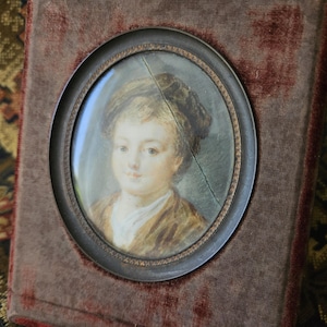 May include: An antique portrait miniature of a person in a brown frame. The oval portrait features a person with light skin, wearing a brown jacket and a white shirt. The portrait is set against a dark red velvet background.