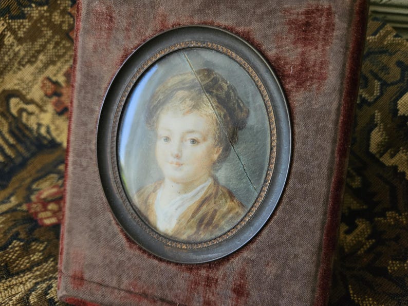 May include: An antique portrait miniature of a person in a brown frame. The oval portrait features a person with light skin, wearing a brown jacket and a white shirt. The portrait is set against a dark red velvet background.