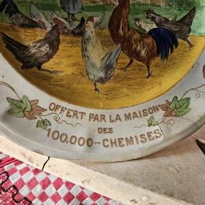 May include: A white ceramic plate with a yellow center and a blue and brown border. The center features a rooster and hens with a leafy design around the edge. The plate has the text "OFFERT PAR LA MAISON DES 100.000 - CHEMISES" printed on the bottom.