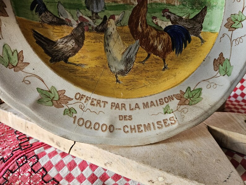 May include: A white ceramic plate with a yellow center and a blue and brown border. The center features a rooster and hens with a leafy design around the edge. The plate has the text "OFFERT PAR LA MAISON DES 100.000 - CHEMISES" printed on the bottom.