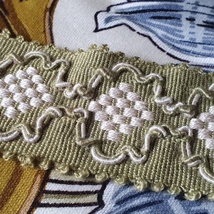 Pode incluir: Close-up de uma guarnição decorativa com uma base tecida verde-oliva e detalhes bordados em branco. A guarnição apresenta um padrão repetitivo de quadrados tecidos e desenhos em laço. O fundo mostra tecido floral em tons de amarelo, azul e branco.