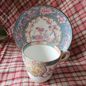 Op de afbeelding: Een vintage theekopje en schotelset met een bloemenpatroon in tinten roze, blauw en groen. Het kopje en de schotel hebben een gouden rand en een delicaat bloemenontwerp.