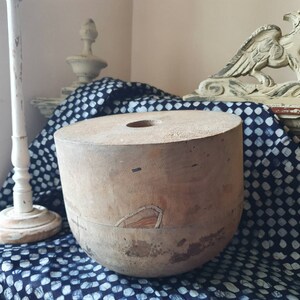 May include: A vintage wooden hat block with a round top and a hole in the center. The block is made of light brown wood and has a worn, rustic appearance.