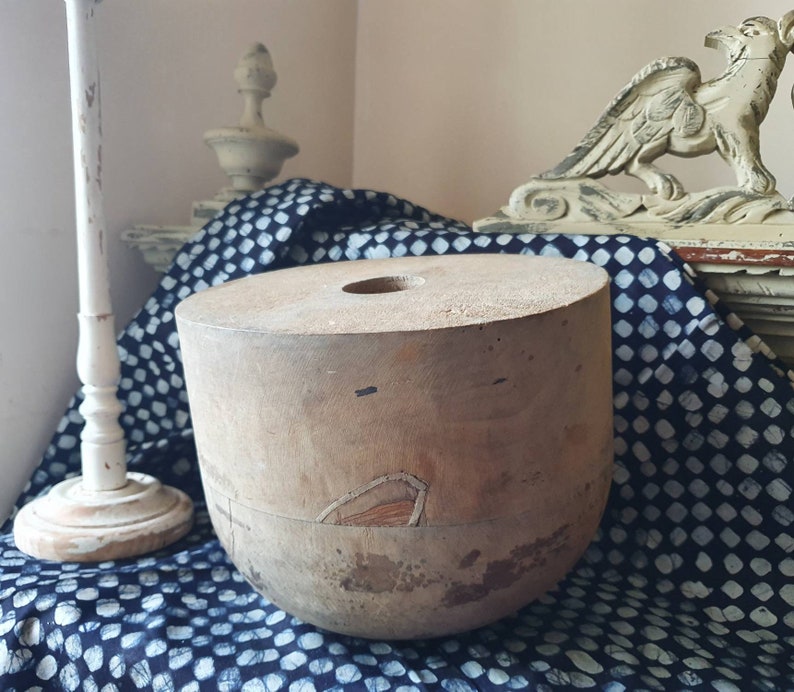 May include: A vintage wooden hat block with a round top and a hole in the center. The block is made of light brown wood and has a worn, rustic appearance.