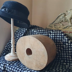 May include: A black hat with a wide brim sits on a white stand. The hat is on a blue and white patterned fabric with a large wooden object in the foreground. The wooden object has a hole in the center.