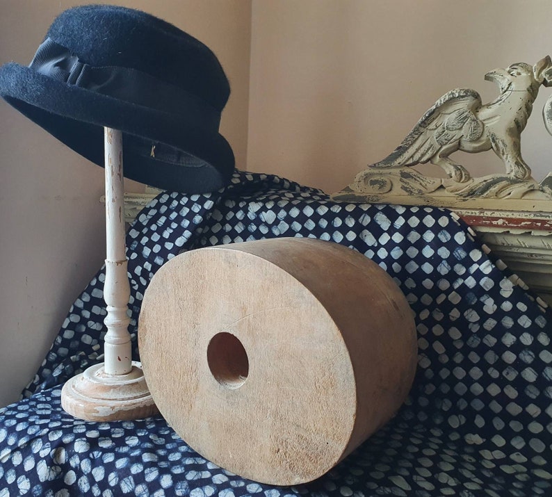 May include: A black hat with a wide brim sits on a white stand. The hat is on a blue and white patterned fabric with a large wooden object in the foreground. The wooden object has a hole in the center.