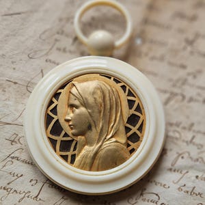 Op de afbeelding: Een witte en gouden hanger met een reliëfbeeld van een vrouw met een sluier. De hanger is bevestigd aan een ring en ligt op een stuk papier met handschrift.