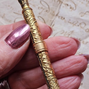 May include: A gold-colored, antique pencil holder with intricate floral and foliate relief carvings. The pencil holder is cylindrical with a small loop at the top and is held in a hand with a pink nail polish.
