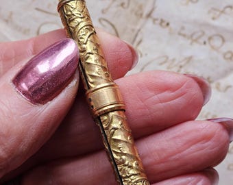 May include: A gold-colored, antique pencil holder with intricate floral and foliate relief carvings. The pencil holder is cylindrical with a small loop at the top and is held in a hand with a pink nail polish.