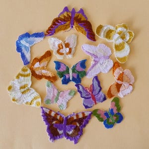 Puede incluir: Una colección de parches de mariposas bordadas de colores, de varios tamaños y diseños. Las mariposas presentan una gama de colores, incluyendo azul, naranja, morado y verde, con costuras detalladas. Están dispuestas sobre un fondo beige claro.
