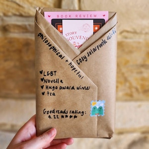 May include: A wrapped package in brown paper, held by a hand. The package contains a book review card and a card with the text "STORY SOUVENIR". Handwritten notes on the package include "philosophical + hopeful", "LGBT", "Novella", and "Hugo award winner". A frog stamp is visible.