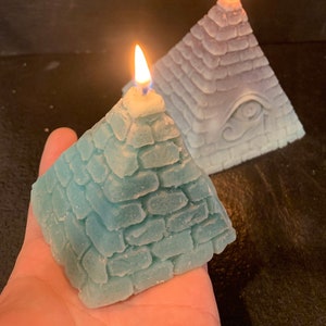 Pode incluir: Duas velas em forma de pirâmide, uma azul e outra roxa, com um design de olho que tudo vê na vela roxa. Ambas as velas estão acesas.