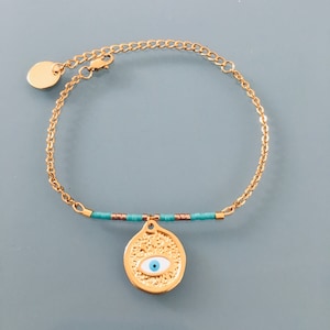 Peut inclure: Un bracelet en chaîne dorée avec un charme œil bleu maléfique et des perles turquoise. Le charme est rond et a une surface texturée.