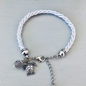 Könnte beinhalten: Weißes Seil-Armband mit silbernen Anhängern, darunter eine Schildkröte, ein Herz mit dem Text "Made with Love" und eine kleine, strukturierte Perle.