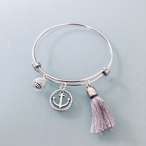 Puede incluir: Un brazalete de plata con un dije de ancla de plata, un dije de espiral de plata y un dije de borla gris.