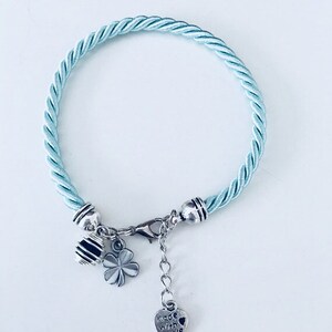 Puede incluir: Un brazalete trenzado azul claro con abalorios plateados, incluyendo un trébol, una abeja y un corazón que dice "Made with Love".