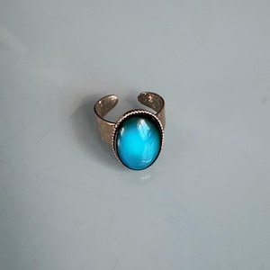 Peut inclure: Bague réglable avec une grosse pierre ovale changeant de couleur. La pierre passe du bleu au turquoise. La bague a un anneau texturé avec un design ouvert.