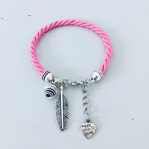 Op de afbeelding: Roze gevlochten koord armband met zilveren bedels, waaronder een veer, een bal en een hartbedel met de tekst "Made with Love".