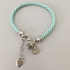 Puede incluir: Un brazalete trenzado azul claro con un dije de plata que dice "Made with Love" y un dije de plata de un Buda sonriente.