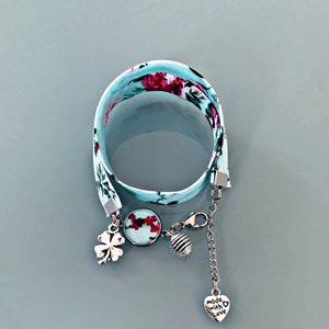 Puede incluir: Un brazalete de tela azul claro con un patrón floral. El brazalete tiene un cierre plateado y varios abalorios, incluyendo un trébol de cuatro hojas, un abalorio redondo con un diseño floral, un abalorio en espiral y un abalorio en forma de corazón que dice "hecho con amor".