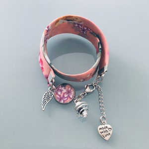 Può includere: Un braccialetto con stampa floreale con sfondo rosa e ciondoli argentati. I ciondoli includono un'ala, un ciondolo rotondo con un design floreale, una perla a forma di gabbia e un ciondolo a cuore con la scritta "made with love".