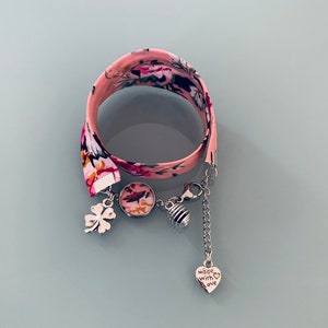 Peut inclure: Un bracelet en tissu floral rose avec des breloques argentées, dont un trèfle, un petit charme rond, un charme en forme de nid d'abeille et un charme en forme de cœur qui dit "fait avec amour".