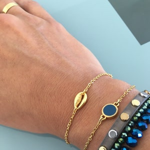 May include: Two gold chain bracelets with a seashell charm and a blue circle charm. The bracelets are worn on a person's wrist.