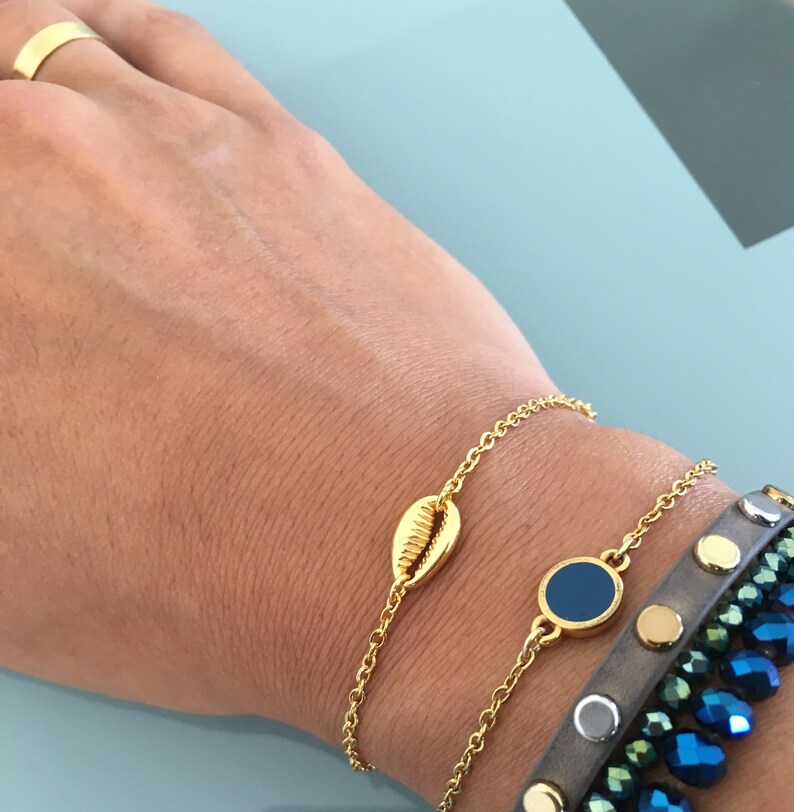 May include: Two gold chain bracelets with a seashell charm and a blue circle charm. The bracelets are worn on a person's wrist.