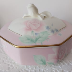 May include: A pink, hexagonal porcelain trinket box with a gold trim. The lid features a white rose-shaped handle and a painted pink rose design with green leaves and blue accents. The box is likely used for storing small items.