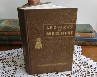 May include: A brown book titled "ABC and XYZ of Bee Culture" with gold lettering and a gold bee emblem. The book is part of "The A. I. Root Bee Library". Other antique books are visible in the background.