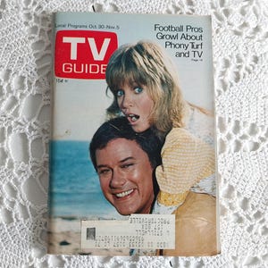May include: A vintage TV Guide magazine cover featuring a couple on a beach. The cover features the text "TV Guide" and "Football Pros Growl About Phony Turf and TV".
