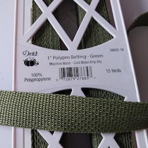 May include: A spool of 1 inch wide green polypro belting by Dritz, made of 100% polypropylene. The white plastic spool holds 15 yards of the woven green strap material and features a barcode, product name, and care instructions printed on it.