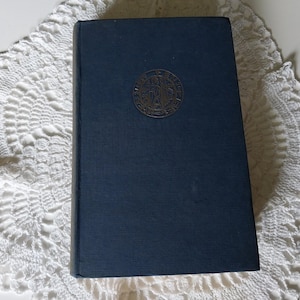 May include: A rectangular hardcover book with a dark blue fabric cover. Centered on the front cover is a circular gold-toned emblem featuring an embossed illustration of a figure holding a staff, surrounded by a decorative border with text.