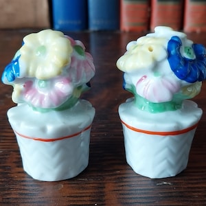 May include: A pair of vintage ceramic salt and pepper shakers. Each shaker is white with a basket-like design and topped with a cluster of colorful flowers in shades of blue, yellow, and pink. The base has an orange trim.