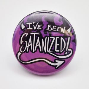 Puede incluir: Un botón redondo morado con el texto "I'VE BEEN SATANIZED!" en letras blancas estilizadas. El fondo presenta un diseño en espiral morado y negro. Una flecha blanca apunta hacia la derecha.